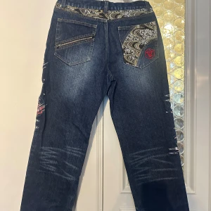 Unika jeans med broderi och tryck - Säljer ett par unika jeans köpta på SEAMS  japan inspirerade med coolt skelett- och blommönster på framsidan. De har även snygga broderade detaljer och en röd patch på baksidan. Jeansen är mörkblå och har en loose fit. Perfekta för dig som vill sticka ut! 🖤82 i midja, innemått längdben 76, passade kille 14 år normallängd. Baggy style. Nypris 2100