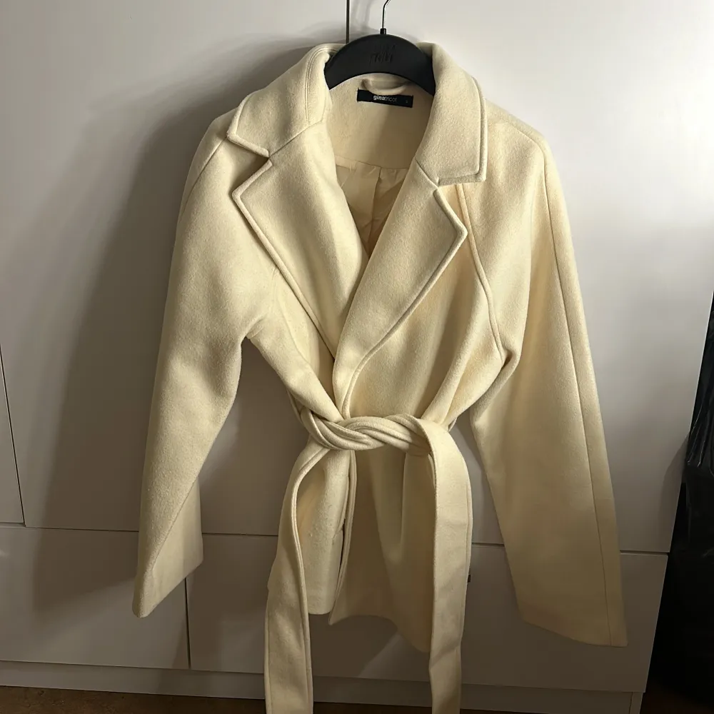 Säljer en superfin beige kappa i ull med knytband i midjan. Den har en klassisk design med långa ärmar och breda slag. Perfekt för höst och vår när du vill vara både stilren och varm. Passar till både vardag och fest!. Jackor.