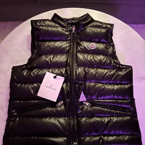 Svart Moncler gui väst - Säljer en snygg svart dunväst från Moncler i nytt skick. Västen har en glansig finish och dragkedja framtill. Perfekt för höst och vinter, och den är både stilren och varm. Passar bra till både vardag och fest. Kom med prisförslag!