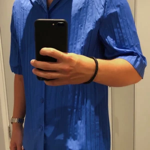 Blå skjorta med korta ärmar - Säljer en snygg blå skjorta med korta ärmar och knappar framtill. Skjortan har ett stilrent vecklat/plisserat material och är perfekt för sommardagar. Den är i bra skick och passar både till vardags och festliga tillfällen. Perfekt för dig som vill ha en avslappnad men ändå stilren look! 💙pris kan alltid diskuteras, skriv om du har frågor