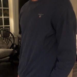 Mörkblå tröja från GANT - Säljer en stilren mörkblå tröja från GANT i mycket bra skick. Tröjan har en liten broderad logga på bröstet och är perfekt för både vardag och lite finare tillfällen. Den är långärmad och gjord i ett mjukt material som känns skönt mot huden. Passar perfekt till jeans eller chinos!