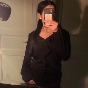 Svart blus med volangdetaljer - Säljer en elegant svart blus med volangdetaljer framtill. Blusen har långa ärmar och en lös passform, perfekt för både vardag och fest. Materialet är mjukt och bekvämt, i storlek M aldrig använd 