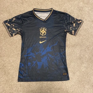 Blå Brasilien fotbollströja från Nike - Säljer en snygg blå Brasilien fotbollströja från Nike. Den har korta ärmar med coola mönster och en gul Nike-logga på bröstet. Tröjan är i bra skick och perfekt för alla fotbollsfans. Den har en V-ringad krage med gul rand och är gjord i ett lätt och bekvämt material.