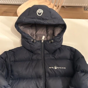 Mörkblå pufferjacka från Sail Racing - Säljer en snygg mörkblå pufferjacka från Sail Racing. Jackan har en dragkedja framtill och är perfekt för kalla dagar. Den är långärmad och har en mysig huva. Perfekt för vintern och hösten! Det är en defekt på armen och en på ryggen inget som är synligt 🧥