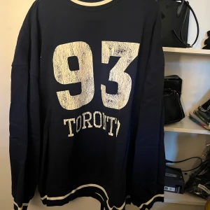 Mörkblå tröja med tryck - Säljer en mörkblå tröja med stort vitt tryck '93 TORONTO' på framsidan. Tröjan har långa ärmar och vita detaljer vid hals, ärmslut och nederkant. Perfekt för en avslappnad stil eller sportig look. Passar bra till jeans eller joggers! Storlek s