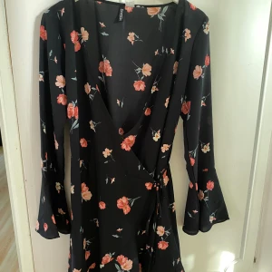 Svart blommig klänning från H&M Divided - Säljer en superfin svart klänning från H&M Divided med blommigt mönster i rött och rosa. Klänningen har en omlott-design och långa ärmar med lätt utsvängda ärmslut. Perfekt för både vardag och fest!