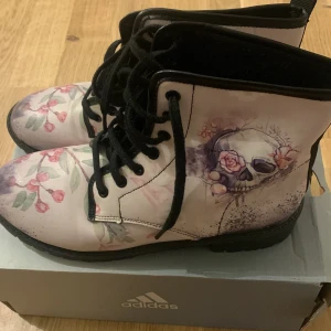 Blommiga boots med dödskallemotiv - Säljer ett par unika boots med blommigt och dödskallemotiv. De har en ljusrosa bas med svarta snören och en cool design. Perfekta för att ge din outfit en edgy touch!