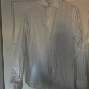 Vit skjorta från J.Lindeberg - Säljer en stilren vit skjorta från J.Lindeberg i slim fit. Perfekt för både vardag och fest! Skjortan har långa ärmar och knappar framtill. Den är i bra skick och passar utmärkt till både jeans och kostymbyxor.
