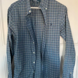 Rutig skjorta från Polo Ralph Lauren - Säljer en snygg rutig skjorta från Polo Ralph Lauren i blå och grå toner. Skjortan är i slim fit och har långa ärmar med knappar. Perfekt för både vardag och festliga tillfällen. Den är i mycket bra skick och redo för en ny ägare!