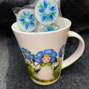 Vit mugg med blå blommor och figur - Charmig vit mugg dekorerad med stora blå blommor och en söt figur i mitten. Perfekt för en mysig kopp te eller kaffe. Muggen har ett bekvämt handtag och är fylld med ljus med matchande blommotiv. En fin presentidé eller ett härligt tillskott till ditt kök. Plus motivet på värmeljus flyter med. 
