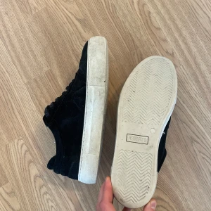 Svarta sneakers i mocka (Arigato) - Säljer ett par svarta sneakers i mocka med vit sula. De har snörning och är i bra skick, perfekt för vardagsbruk. Skorna har en stilren design och passar till det mesta. Storleken är 42.
