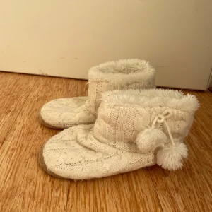 Vita stickade tofflor med pom-poms - Supermysiga vita stickade tofflor med fluffigt foder och söta pom-poms på sidan. Perfekta för att hålla fötterna varma under kalla vinterdagar. De har en mjuk och bekväm passform som gör dem idealiska för hemmabruk. Ett måste för mysiga kvällar!
