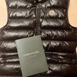 Svart dunväst från Moncler - Säljer en snygg svart dunväst från Moncler i mycket bra skick. Västen har en glansig finish och en praktisk dragkedja framtill. Perfekt för höst och vinter, och den är både stilren och varm. Passar bra till både vardag och festliga tillfällen.