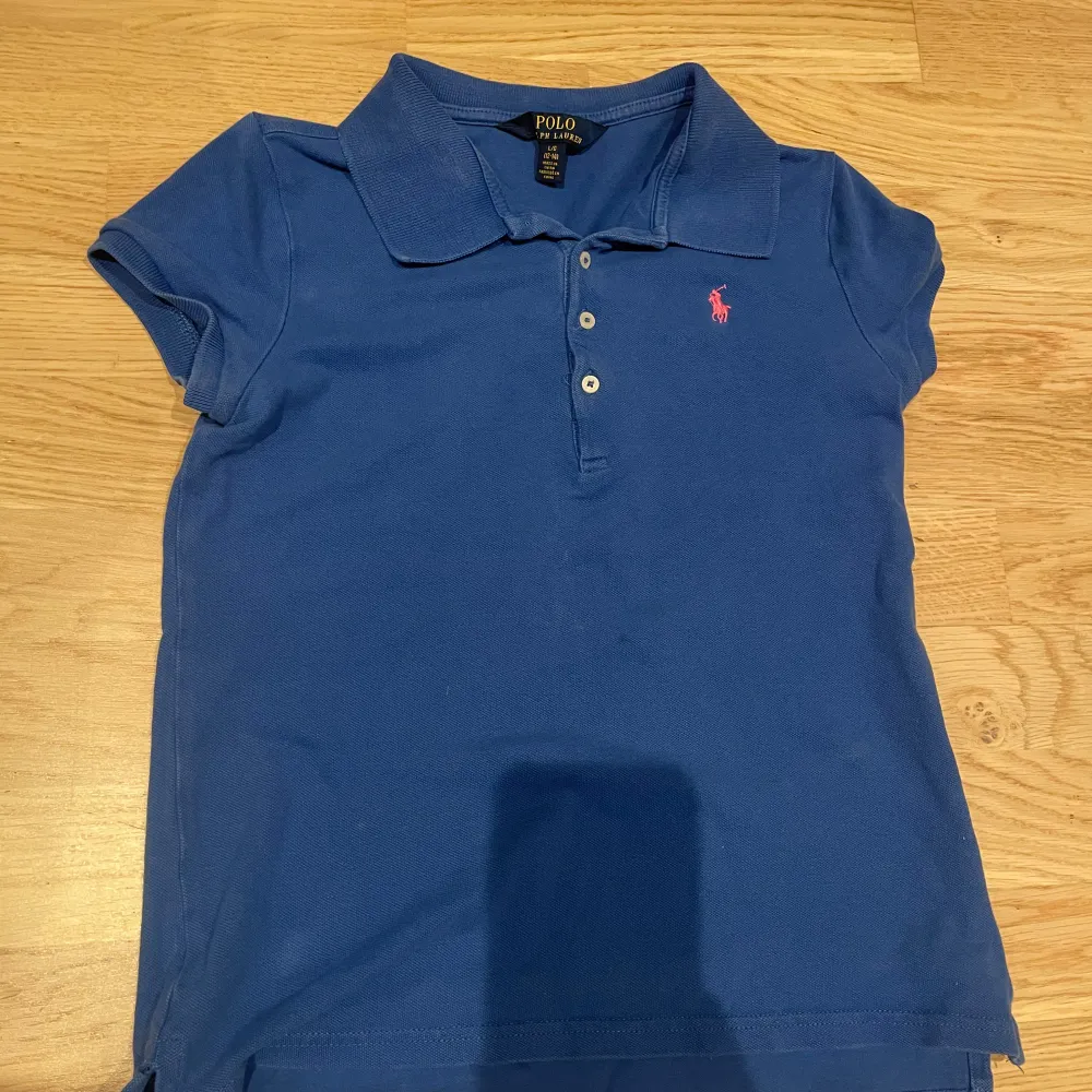 Säljer en klassisk blå pikétröja från Ralph Lauren i storlek 12-14. Tröjan har korta ärmar och en liten rosa logga på bröstet. Perfekt för en avslappnad men stilren look. Använd men i bra skick.. T-shirts.