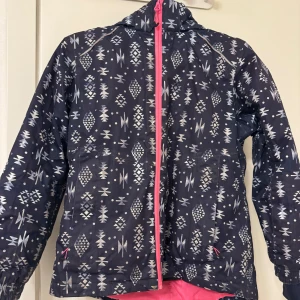 Mönstrad vindjacka med dragkedja - Säljer en cool mönstrad barn- vindjacka i mörkblått med vita geometriska mönster. Jackan har en rosa dragkedja och är perfekt för höst och vår. Den har långa ärmar och en praktisk huva. Perfekt för att hålla sig varm och stilren i blåsigt väder