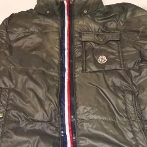 Grön dunjacka från Moncler - Säljer en snygg grön dunjacka från Moncler. Jackan har en cool röd, vit och blå rand längs dragkedjan och en praktisk bröstficka med knapp. Perfekt för kyliga dagar och håller dig varm med sin dunfyllning. Passar både till vardags och lite finare tillfällen. 🧥 lite förstörd endast luvan är borta det är ända problemet