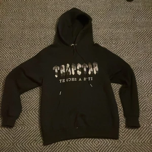 Svart hoodie från Trapstar - Säljer en svart hoodie från Trapstar i storlek S. Den har en cool text på framsidan och är perfekt för både höst och vinter. Hoodien är i bra skick och superbekväm att ha på sig. Perfekt för en avslappnad stil eller streetwear-look!