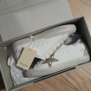 Golden Goose sneakers med glittrig stjärna - Säljer ett par snygga Golden Goose sneakers i vitt skinn med en glittrig stjärna på sidan. Skorna har silverdetaljer på hälen och klassisk snörning. Perfekta för att ge din outfit en extra touch av stil! De är i nytt skick och kommer med originalförpackning.