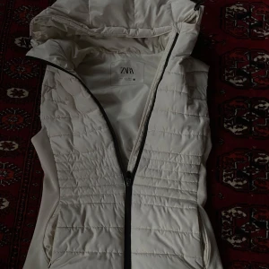 Vit quiltad väst från Zara - Säljer en snygg vit quiltad väst från Zara. Den har en dragkedja framtill och en mysig huva. Perfekt för höst och vår när du vill hålla dig varm men ändå se stilren ut. Den är som ny och knappast andvänd heller. Storlek s