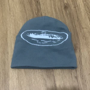 Grå mössa med tryck - Säljer en snygg Cortiez  grå  mössa med ett coolt vitt tryck på framsidan. Perfekt för att hålla sig varm under kyliga dagar. Den är mjuk och bekväm, idealisk för både vardag och utomhusaktiviteter. Passar de flesta huvudstorlekar.