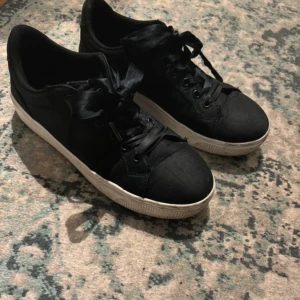 Svarta sneakers med snörning - Säljer ett par svarta sneakers i bra skick. De har en klassisk design med snörning och en vit sula som ger en snygg kontrast. Perfekta för vardagsbruk och passar till det mesta i garderoben. Storleken är dam 37. Står emot lätt regn så helt perfekt för lite rusigare dagar! 