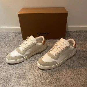 Burberry skor - Säljer ett par snygga vita sneakers från Burberry i mycket bra skick. De har en klassisk design med beiga detalger och den ikoniska Burberry-rutiga baksidan. Perfekta för både vardag och fest! Storlek är 42,5 men passar även 42.