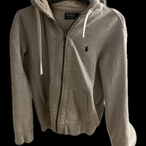 Grå hoodie från Polo Ralph Lauren - Säljer en snygg grå hoodie från Polo Ralph Lauren. Den har en dragkedja framtill och klassisk logga på bröstet. Perfekt för höst och vår, med ribbade muddar och en skön passform. Perfekt för en casual look eller mysiga dagar hemma. Lite sliten vid ena armen! Därav priset !