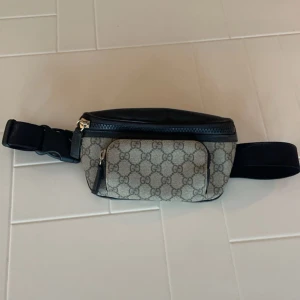 Gucci magväska med monogram - Säljer en snygg Gucci magväska i mycket bra skick. Den har det klassiska GG-monogrammet i beige och svart med svarta läderdetaljer. Väskan har en justerbar rem och dragkedja både fram och bak. Perfekt för att hålla dina saker säkra och stiliga på språng!