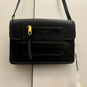 Svart axelväska från Marc Jacobs - Snygg svart axelväska från Marc Jacobs i skinn med gulddetaljer. Väskan har en justerbar axelrem och en praktisk ficka framtill med dragkedja. Perfekt för både vardag och fest!