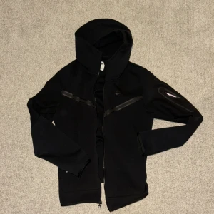Svart Nike Tech Fleece hoodie  - Svart TechFleece hoodie i storleken S. Säljer den då den inte kom till användning. Den är gjord av främst polyester men även lite bomull. Obs att dragkedjan på tröjan/hoodien ej fungerar. 