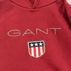 Röd hoodie från GANT - Säljer en snygg röd hoodie från GANT med deras klassiska logga och ett coolt emblem med stjärnor och ränder på bröstet. Perfekt för en avslappnad stil och passar bra till både jeans och joggers. Den är i bra skick och superbekväm att ha på sig. Perfekt för höst och vinter!   Storlek 158/164 cm