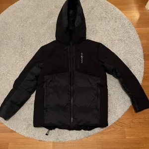 Svart dunjacka från Sail racing  - Säljer en snygg svart dunjacka från Sail racing . Den är perfekt för kalla vinterdagar med sin varma vaddering och huva. Jackan har en dragkedja framtill och flera praktiska fickor. Den är endast använd ett fåtal gånger och i perfekt nyskick!