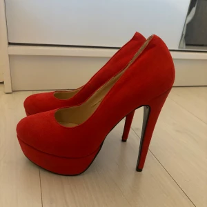 Röda pumps med hög klack - Säljer ett par snygga röda pumps i mycket bra skick. De har en hög klack och en platåsula som ger extra höjd och stil. Perfekta för festliga tillfällen eller en utekväll. Materialet ser ut att vara mocka, vilket ger en lyxig känsla.