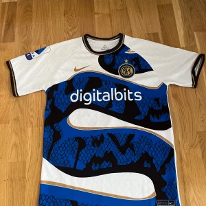 Inter Milan fotbollströja - Säljer en snygg Inter Milan fotbollströja från Nike. Den är vit med blå och svarta mönster och har korta ärmar. Tröjan har klubbens emblem på bröstet.