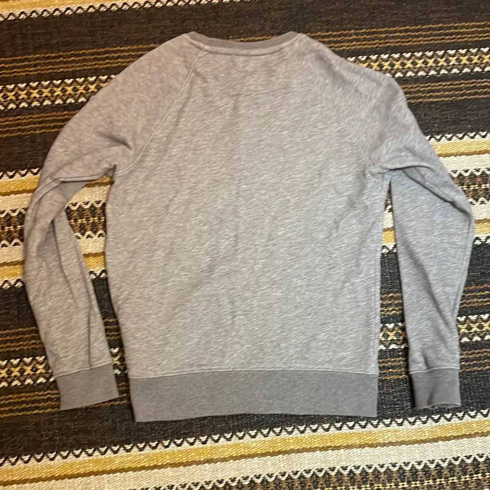 Säljer en snygg grå sweatshirt från GANT i mycket bra skick. Den har ett coolt emblem med stjärnor och ränder på bröstet och GANT-loggan broderad ovanför. Perfekt för en avslappnad stil och passar bra till både jeans och chinos. Superbekväm och stilren!. Tröjor & Koftor.