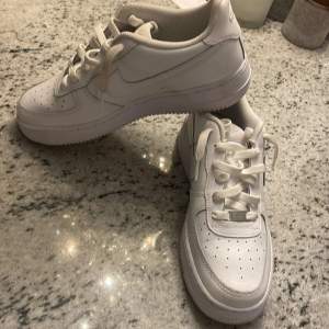 Säljer ett par klassiska vita Nike Air Force sneakers i mycket bra skick. De har en stilren design med snörning och är perfekta för både vardag och fest. Skorna är i skinn och har den ikoniska Nike-loggan på sidan och hälen. Har använts 1 gång och är helt nya