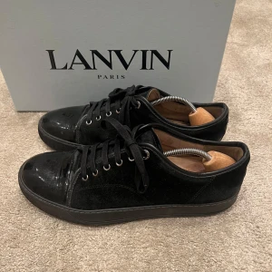 Svarta sneakers från Lanvin - Säljer ett par stilrena svarta sneakers från Lanvin i använt skick. Ett hål rakt genom sulan på ena skon, går att laga hos skomakare, andra defekter så som vattenskador på mockan och fläckar, men mycket går att rengöra. Skorna har en snygg kombination av skinn och mocka med klassisk snörning. Perfekta för både vardag och fest. Originalförpackning, dustbags, extra snören och kvitto medföljer. Nypris: 5200kr. Fraktar på köparens bekostnad samma dag som beställning🤝