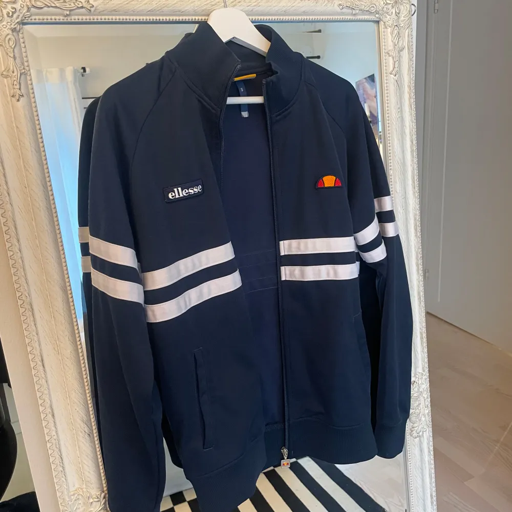 Säljer en snygg mörkblå jacka från Ellesse med vita ränder över bröstet och ärmarna. Jackan har en dragkedja framtill och hög krage. Perfekt för en sportig look eller en avslappnad dag ute!. Hoodies.