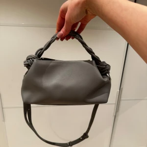 Grå axelväska med flätat handtag - Säljer en snygg grå axelväska med ett unikt flätat handtag. Väskan är i mycket bra skick och perfekt för både vardag och fest. Den har en justerbar axelrem💕 Passar perfekt till alla outfits!💕