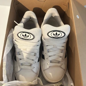 Adidas campus  sneakers i vitt och svart - Säljer ett par snygga Adidas sneakers i vitt med svarta detaljer. De har den klassiska tre-randiga designen och är i mycket bra skick. Perfekta för både vardag och träning. Skostorlek dam 38.