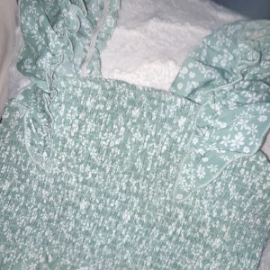 Grön blommig topp med volangdetaljer - Söt grön topp med blommigt mönster och volangdetaljer på axlarna. Perfekt för sommardagar eller en avslappnad vårlook. Toppen har en smockad design som ger en bekväm och flexibel passform.