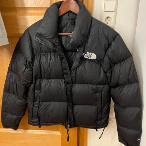 Svart dunjacka från The North Face - Säljer en snygg och varm svart dunjacka från The North Face. Perfekt för kalla vinterdagar. Jackan har en dragkedja framtill och en hög krage för extra värme. Den är i mycket bra skick och har en klassisk pufferstil. Passar perfekt för både vardag och utomhusaktiviteter.