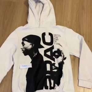 Vit hoodie med 2Pac-tryck - Säljer en vit hoodie med stort 2Pac-tryck på framsidan och låtlista på baksidan. Perfekt för alla hiphop-fans! Den är i bra skick och har en skön passform. Huvan är mysig och perfekt för kyliga dagar. Passar både till vardags och chillkvällar.