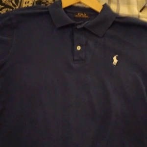 Blå pikétröja från Ralph Lauren - Säljer en klassisk blå pikétröja från Ralph Lauren. Tröjan har en broderad logga på bröstet och knappar framtill. Perfekt för en stilren och avslappnad look.