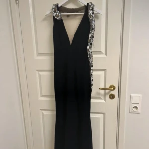 Svart långklänning - Svart långklänning med genomskinlig rygg och v-ringning. Från höften och upp runt halsen går en ”orm” av stenar. Dubbelt tyg och insydda kupor.