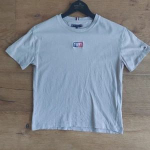 Beige t-shirt från Tommy Hilfiger - Säljer en snygg beige t-shirt från Tommy Hilfiger. Den har en klassisk rund hals och korta ärmar. På framsidan finns en broderad Tommy-logga i rött, vitt och blått. Perfekt för en avslappnad stil! OPS har två små hål på framsida  och ett på baksidan.