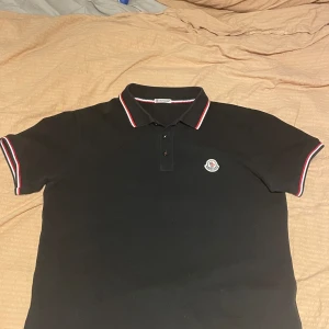 Svart kortärmad pikétröja från Moncler - Säljer en snygg svart pikétröja 1: 1. Den har korta ärmar och en klassisk krage med röd och vit rand. Moncler-loggan är broderad på bröstet. Perfekt för en stilren och avslappnad look.