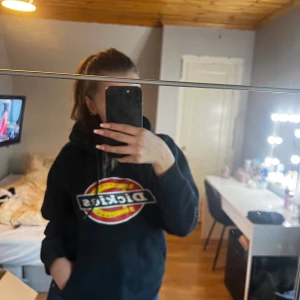 Svart hoodie från Dickies - Säljer en svart hoodie från Dickies med deras ikoniska logga i rött och gult på bröstet. Den är superbekväm och perfekt för både chill och vardag. Har en stor ficka fram och en mysig huva. Perfekt för höst och vinter!