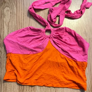 Färgglad topp med knytband - Säljer en superfin och färgglad topp i rosa och orange. Den har ett unikt knytband runt halsen och en cool design med en ringdetalj framtill. 