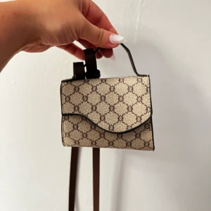 Mönstrad axelväska - Säljer en superfin liten axelväska/walletbag med ett klassiskt mönster i beige och brunt. Väskan har en justerbar rem och stängs med en tryckknapp. 